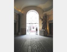 Interno di un portico monumentale con pavimento in pietra e una corsia centrale in ciottoli che conduce a un grande arco d’ingresso illuminato verso l’esterno. A destra si trova una grande statua su piedistallo accanto a colonne e pareti con modanature classiche.