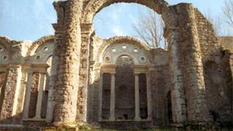 Rovine in pietra di un edificio storico con grandi arcate crollate e colonne ancora in piedi, con pareti traforate da finestre ad arco. In primo piano un prato e sullo sfondo cielo aperto e alberi spogli.