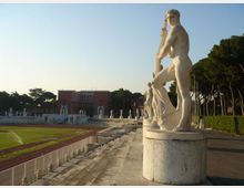 Viale di uno stadio all’aperto con gradinate in pietra e pista di atletica, fiancheggiato da una fila di statue classiche in marmo su basamenti. Sullo sfondo si vedono un edificio in mattoni rossi e alberi alti lungo il perimetro.
