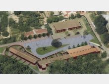 Veduta aerea di un complesso rurale con più edifici rettangolari dai tetti rossi disposti attorno a una grande area pavimentata. Al centro ci sono aiuole verdi con alberi e vialetti curvi, circondati da campi e vegetazione.