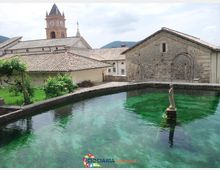 Vasca d’acqua limpida color verde smeraldo in un borgo storico, circondata da edifici in pietra e tetti in coppi. Sullo sfondo si vede un campanile con orologio e colline basse sotto un cielo nuvoloso.