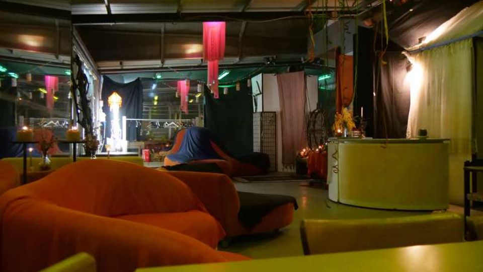 Interno di un locale tipo lounge con soffitto industriale e illuminazione soffusa, arredato con grandi divani e pouf arancioni. Nello spazio si vedono tendaggi colorati e pannelli divisori, con un bancone chiaro sulla destra e luci decorative sullo sfondo.