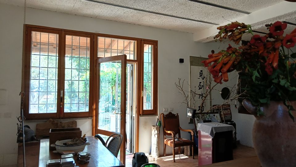 Interno di una sala da pranzo rustica con soffitto a travi, grande tavolo in legno e ampie finestre e porta finestra con griglie che si aprono su un patio esterno. Arredi e oggetti decorativi sono distribuiti lungo le pareti, con un grande vaso di fiori in primo piano.