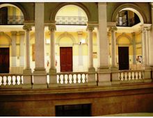 Cortile interno di un edificio storico con porticato su due livelli, grandi archi e colonne in pietra. Al piano superiore si vedono balaustre e ringhiere in ferro, con pareti color crema e porte scure sullo sfondo.