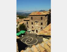 Piazza lastricata in pietra con disegno circolare, circondata da edifici in muratura con tetti in coppi; in primo piano un grande palazzo in pietra con persiane scure e portale ad arco. Sullo sfondo si vedono colline e montagne sotto un cielo sereno.