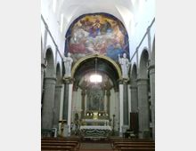 Interno di una chiesa con navata centrale fiancheggiata da arcate e colonne in pietra, che conduce all’altare maggiore rialzato. Sopra l’altare si vede un grande affresco semicircolare e statue su piedistalli ai lati dell’arco, con banchi in legno in primo piano.