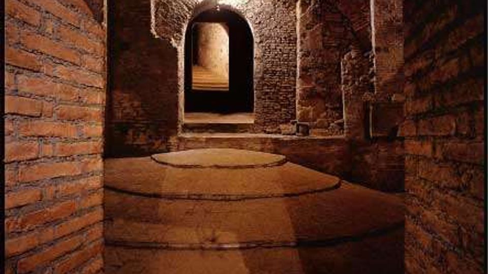 Corridoio sotterraneo in mattoni e pietra con soffitto a volta e una serie di archi, illuminato da una lampada a parete. Pavimento in pietra con gradini circolari e passaggio che conduce a un’apertura ad arco in fondo.