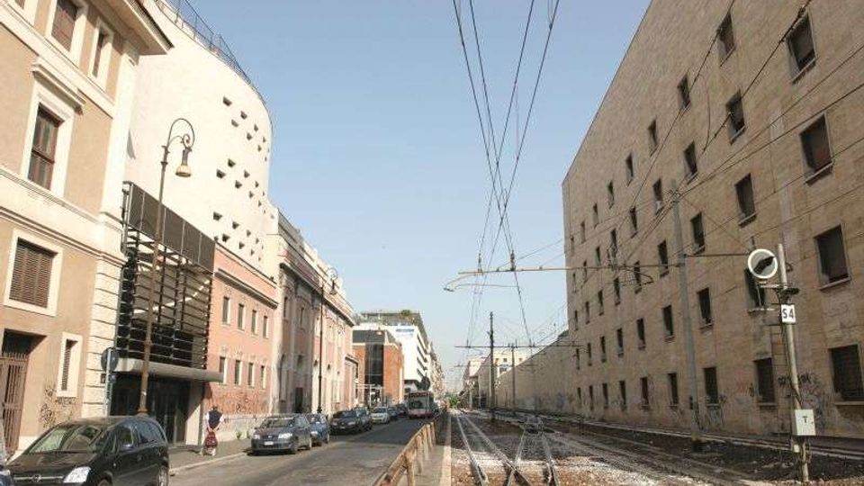 Strada urbana rettilinea con binari ferroviari e cavi aerei sul lato destro, affiancata da un lungo edificio in mattoni con piccole finestre. Sul lato sinistro si vedono palazzi moderni e storici, auto parcheggiate e lampioni lungo il marciapiede.