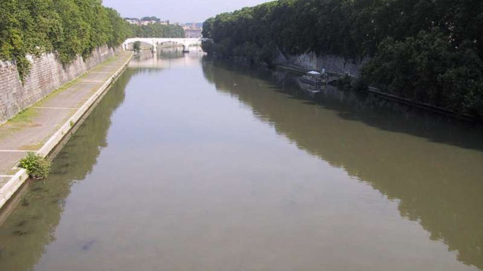 Veduta di un fiume o canale urbano con acqua calma, fiancheggiato da alti argini in muratura e da una lunga banchina pedonale sul lato sinistro. Entrambi i lati sono bordati da alberi fitti e sullo sfondo si vede un ponte ad arcate in pietra.
