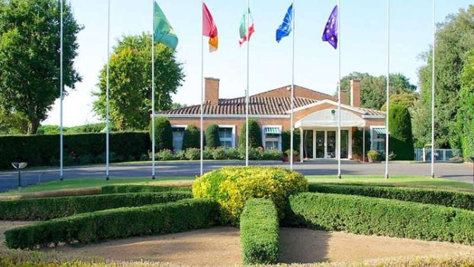Edificio basso in mattoni con tetto a falde e ingresso porticato con colonne, situato dietro una rotonda con aiuole e siepi geometriche. Davanti alla facciata sono presenti vari pennoni con bandiere, circondati da alberi e verde curato.