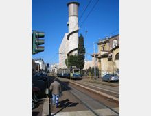 Strada urbana con binari del tram e linea aerea, affiancata da edifici e auto parcheggiate. Sullo sfondo si vede una torre cilindrica in cemento con una rampa elicoidale esterna e un tram che percorre la carreggiata.