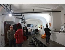 Interno di uno spazio tipo studio o piccola galleria con soffitto a volta in mattoni dipinti di bianco e tubazioni a vista, illuminato da luci da set. Lungo la sala ci sono sedie pieghevoli, attrezzatura video e fotografica, schermi e quadri appesi alle pareti, con pavimento in legno scuro.