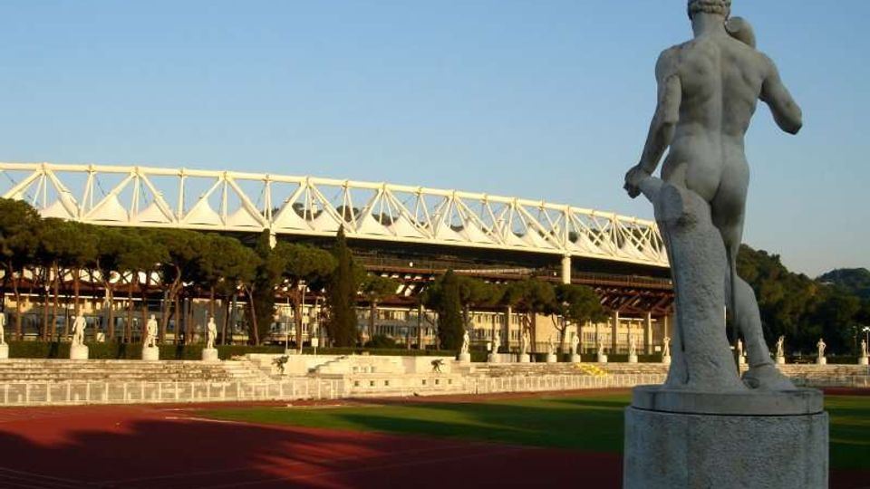 Statua in pietra su piedistallo accanto a una pista di atletica, con sullo sfondo un grande stadio con struttura bianca a traliccio e alberi allineati lungo le gradinate sotto un cielo sereno.