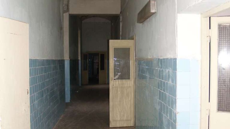 Corridoio interno lungo e stretto con soffitto a volta, pareti intonacate chiare e rivestimento di piastrelle azzurre nella parte bassa. Diverse porte laterali, una porta in legno con griglia semiaperta e pavimento in cemento con segni di usura.