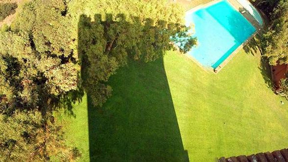 Vista dall’alto di un’area verde con prato curato, circondata da alberi, con una piscina rettangolare azzurra nell’angolo superiore destro. Una grande ombra di un edificio si estende sul prato e si intravede un piccolo tetto in tegole sul lato destro.
