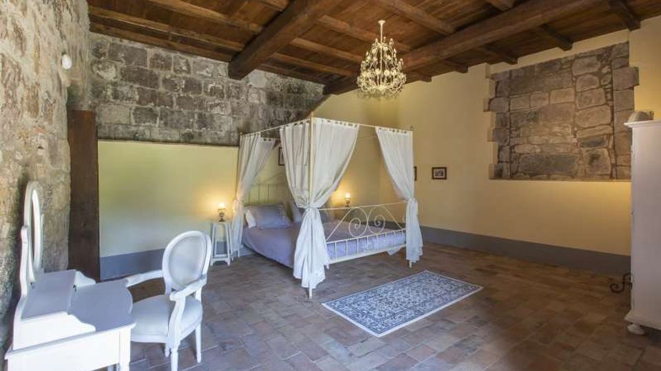 Camera da letto in stile rustico con soffitto a travi in legno, pareti in pietra a vista e pavimento in cotto. Al centro c’è un letto a baldacchino bianco con tende, illuminato da abat-jour e da un lampadario, con una piccola scrivania e sedia bianche sul lato.
