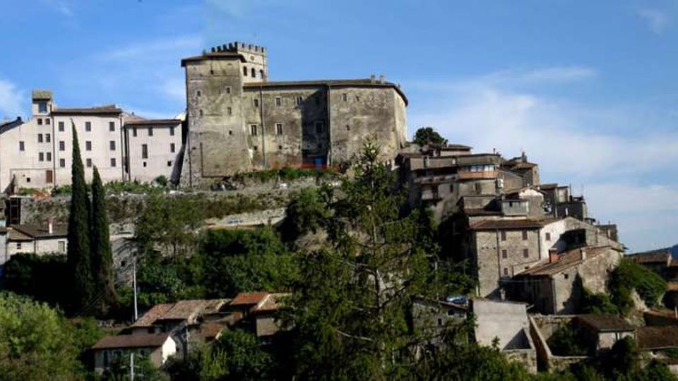 Veduta panoramica di un borgo su collina con un grande castello in pietra e torre quadrata in cima, circondato da edifici storici compatti con tetti in coppi. In primo piano si vedono alberi e vegetazione, con il profilo delle colline sullo sfondo sotto un cielo sereno.