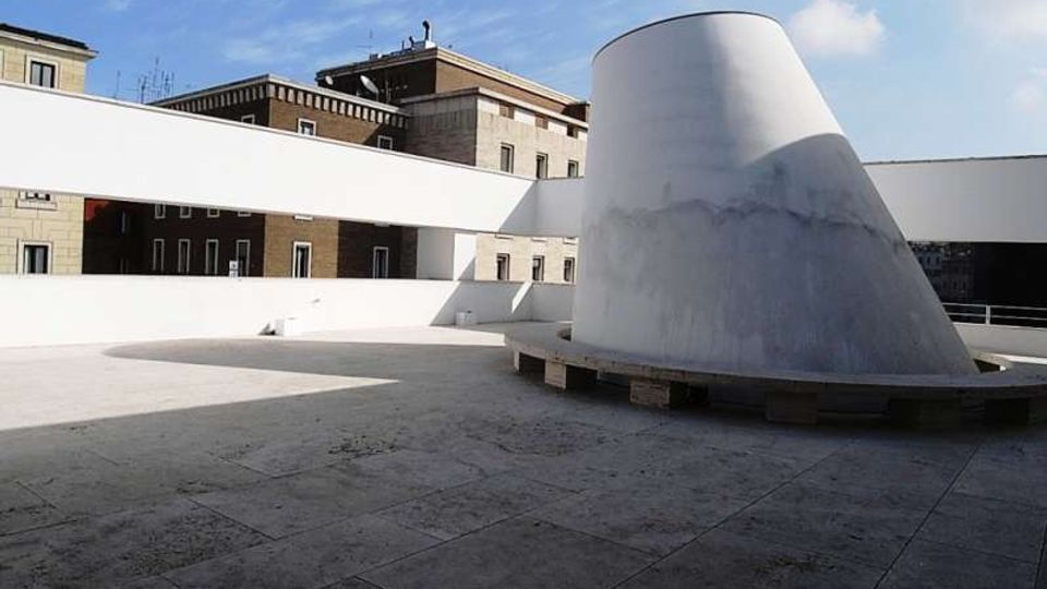 Terrazza pavimentata con ampi parapetti bianchi e una grande struttura conica bianca al centro. Sullo sfondo si vedono edifici urbani in muratura sotto un cielo azzurro.