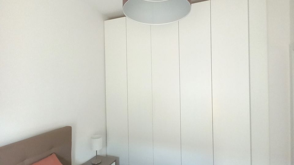 Camera da letto moderna con letto matrimoniale in primo piano, comodino con lampada e grande armadio bianco a pannelli lungo la parete. Soffitto bianco con lampadario a sospensione cilindrico e pavimento in legno scuro.