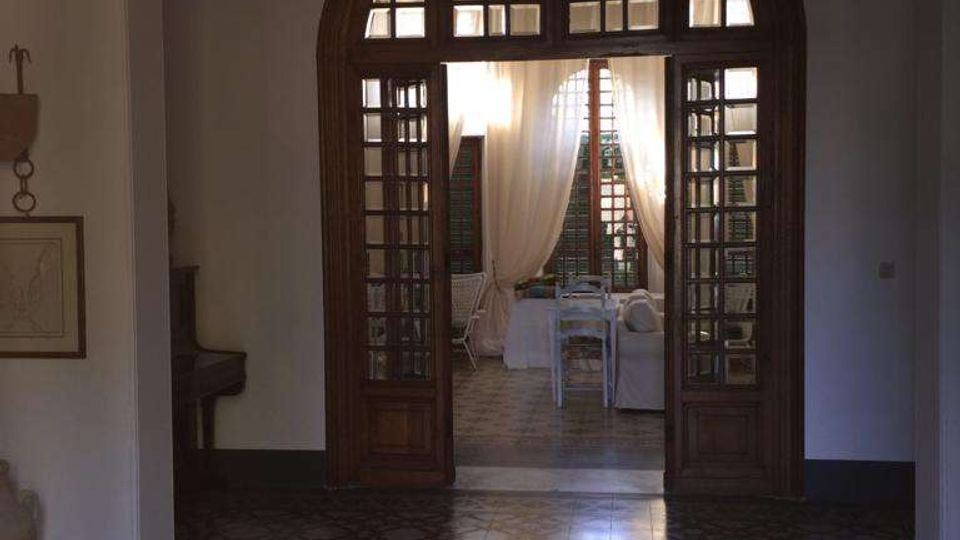 Interno di un edificio con grande arco e portale in legno a vetrate che introduce a una stanza luminosa con tende bianche e arredi chiari. Pavimento in piastrelle decorative con motivi geometrici e pareti intonacate chiare.