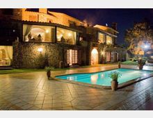 Villa in pietra illuminata di notte con ampie vetrate e porticati, affacciata su una piscina rettangolare con acqua turchese. Intorno alla piscina c’è una grande pavimentazione in lastre e vasi con piante, con giardino e alberi sullo sfondo.