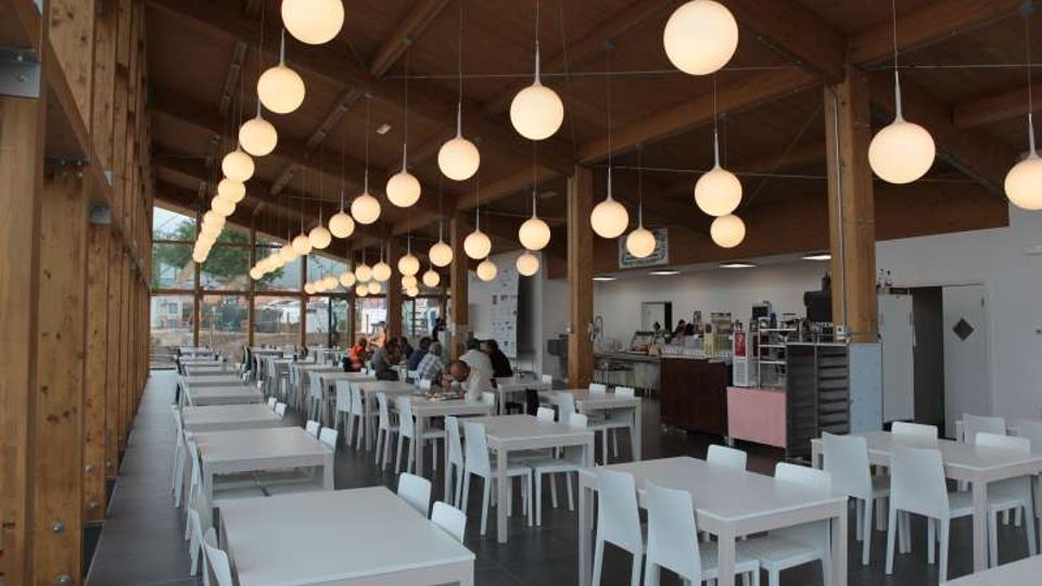 Ampia sala mensa moderna con struttura in legno a vista, grandi vetrate laterali e file di lampade sferiche sospese dal soffitto. Numerosi tavoli e sedie bianchi sono disposti in file, con un’area self-service e bancone sul fondo.
