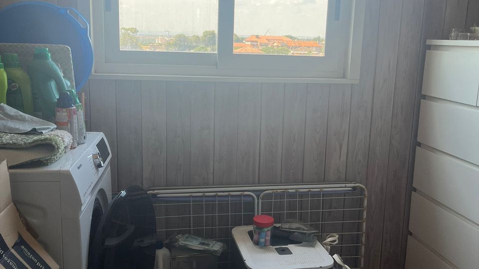 Piccolo locale lavanderia con pareti rivestite in legno e finestra scorrevole centrale che affaccia sui tetti e sul cielo. In primo piano si vedono una lavatrice, detersivi e contenitori, un tappetino a terra e una cassettiera bianca sul lato destro.