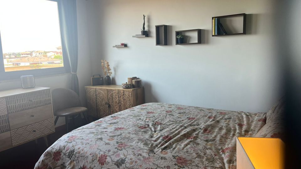 Camera da letto luminosa con letto matrimoniale e copriletto floreale, parete bianca con mensole a cubo e soffitto con travi scure. Una grande finestra con tenda grigia illumina la stanza, con cassettiera e mobile decorato vicino alla parete.