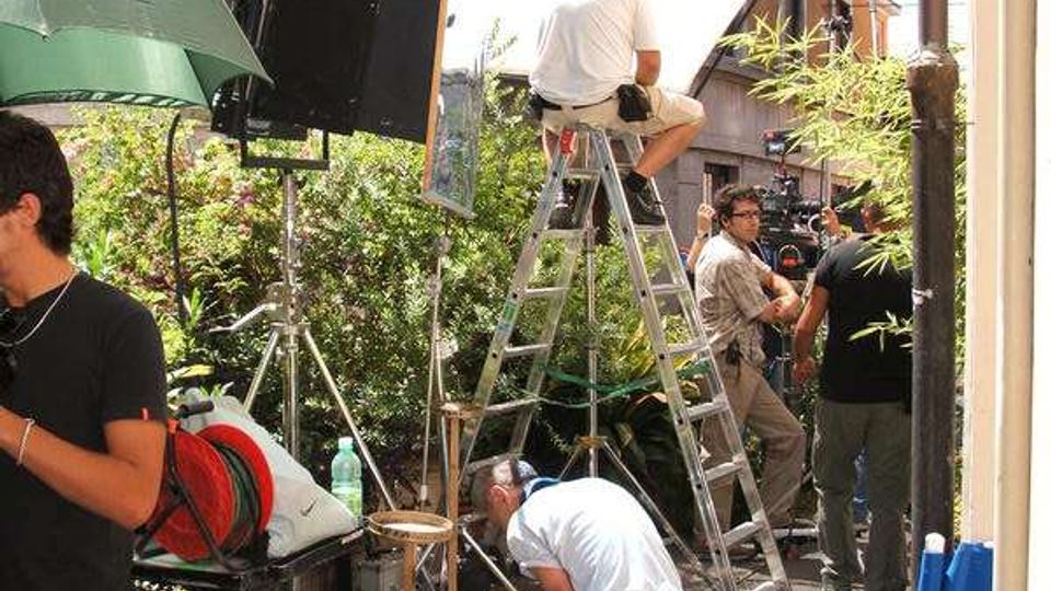 Set cinematografico all’aperto in una strada urbana, con grandi pannelli riflettenti e luci su cavalletti, scale a pioli e cavi elettrici sul pavimento. Sullo sfondo si vedono facciate di edifici e vegetazione lungo il bordo della strada.