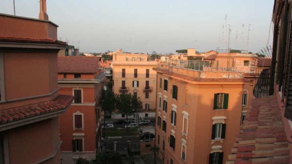 Veduta dall’alto su un quartiere urbano con palazzi residenziali color ocra e rosa, finestre con persiane verdi e tetti in tegole rosse. Tra gli edifici si intravede una strada alberata e, sullo sfondo, altri tetti con antenne sotto un cielo sereno.