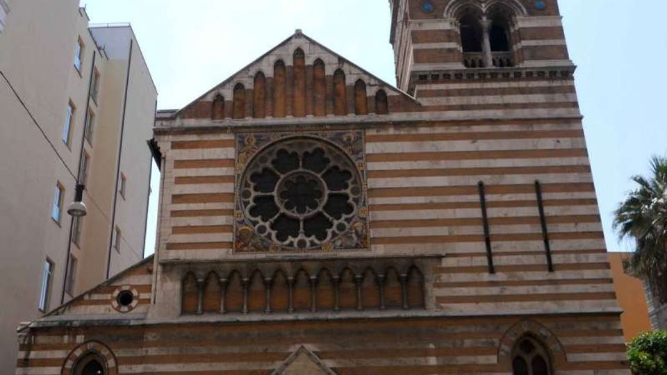 Facciata di una chiesa in stile romanico-gotico con pietra a fasce orizzontali, grande rosone centrale e portale ad arco ogivale. Accanto si innalza un campanile alto con logge ad archi sovrapposti, in un contesto urbano tra edifici moderni.
