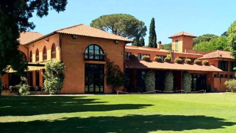 Edificio in stile villa mediterranea con facciata color terracotta e tetti in coppi, caratterizzato da grandi finestre ad arco e porticati con colonne. Davanti si estende un ampio prato curato, con alberi alti e vegetazione sullo sfondo.