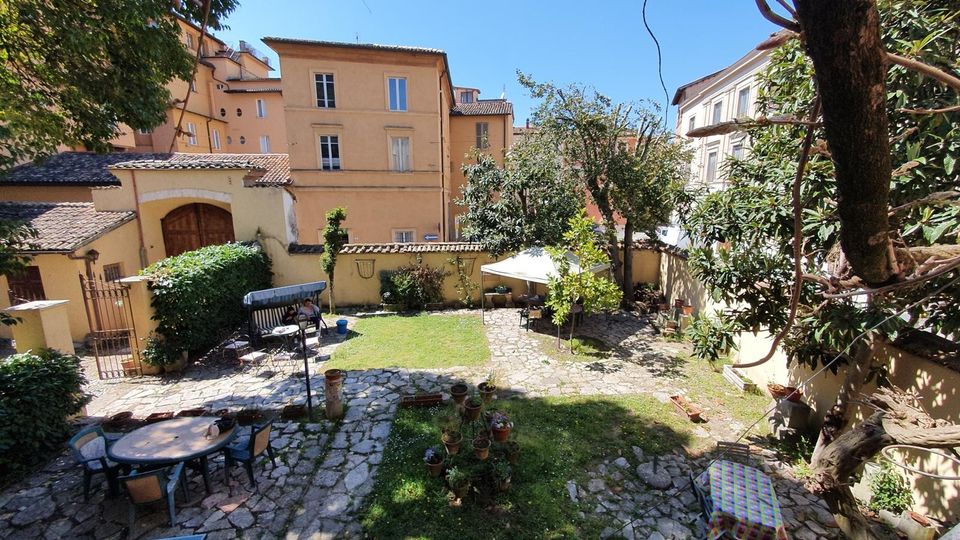 Cortile interno con area verde e pavimentazione in pietra, delimitato da muri e circondato da edifici in intonaco chiaro. Sono presenti alberi e siepi, tavoli e sedie da esterno, vasi con piante e un gazebo/ombrellone su una zona lastricata.