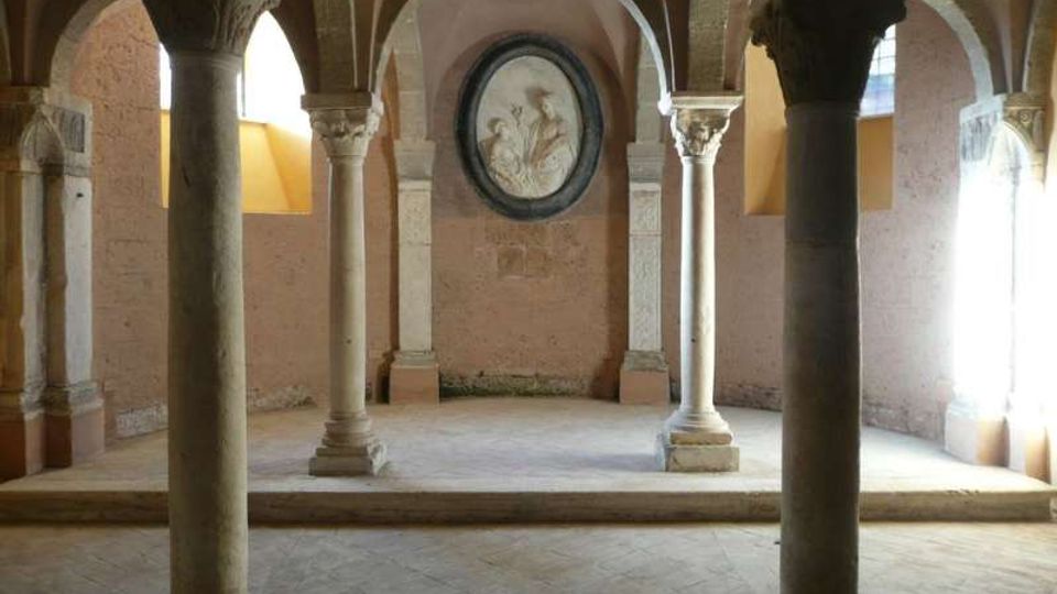 Interno di una sala in pietra con colonne massicce e archi a volta che sostengono il soffitto. Sul fondo, una parete semicircolare ospita un medaglione decorativo, con luce naturale che entra da aperture laterali.