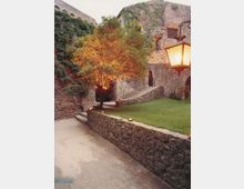 Cortile in pietra di un edificio storico con muri antichi coperti di vegetazione, una scalinata che conduce a un arco e un prato curato con un albero illuminato. In primo piano si vede un vialetto con muretto in pietra e un lampione in stile classico.