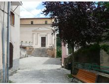 Stradina in salita di un borgo che conduce a un edificio con facciata classica, portale centrale e ampia scalinata in pietra. Ai lati ci sono case addossate, un grande albero che ombreggia il percorso, ringhiere e una panchina lungo il muro.