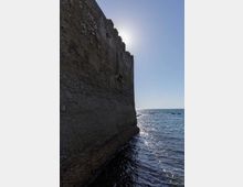 Vista laterale di una massiccia fortificazione in pietra con merlature, affacciata direttamente sul mare. L’acqua lambisce la base del muro mentre il sole alto illumina l’orizzonte marino.