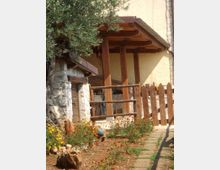 Portico in legno addossato a una casa con pareti intonacate, sostenuto da pilastri e coperto da una piccola tettoia spiovente. Davanti si vedono una staccionata rustica, un’aiuola con fiori e un vialetto di pietra accanto a un albero.