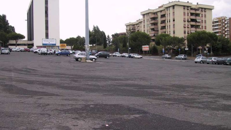 Ampio parcheggio asfaltato quasi vuoto, con diverse auto parcheggiate e un alto palo della luce al centro. Sullo sfondo si vedono un edificio moderno a torre bianco e vari palazzi residenziali con balconi, circondati da alberi lungo la strada.