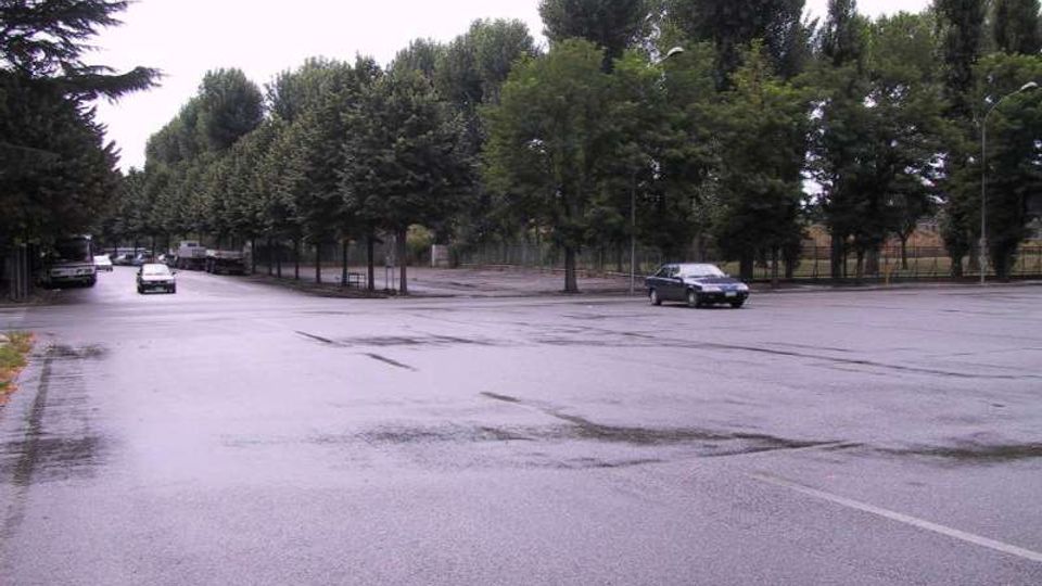 Ampia strada asfaltata con carreggiate larghe e segnaletica poco visibile, fiancheggiata da filari di alberi alti e densi. Sullo sfondo si vedono recinzioni e lampioni lungo il bordo della strada.