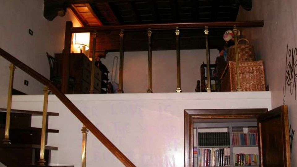 Interno di una casa con soppalco in legno e parapetto con colonnine, raggiungibile tramite una scala in legno con corrimano. In basso si vede una porta aperta su una libreria a muro, con pareti chiare e soffitto con travi scure.