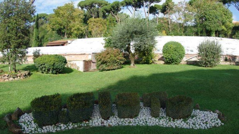 Giardino curato con ampio prato verde, siepi potate e aiuole bordate di ciottoli bianchi in primo piano. Sullo sfondo si vede una lunga struttura bassa con copertura bianca, circondata da alberi e pini marittimi.
