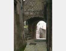 Vicolo stretto in un centro storico, con pavimentazione in ciottoli e un arco in pietra grezza che collega due edifici. A sinistra ci sono gradini in pietra con corrimano, mentre oltre l’arco si intravede una strada in discesa con case e scalinate.