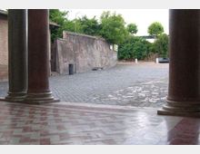Portico con grandi colonne in pietra e pavimento a piastrelle, affacciato su una piazzetta lastricata e un’area pavimentata in pietra. Sullo sfondo si vede un lungo muro in intonaco, un lampione e vegetazione alberata lungo il perimetro.