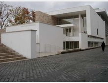 Edificio moderno bianco con volumi geometrici, grandi vetrate e pilastri, affiancato da un muro in pietra. Davanti si vede una strada in ciottoli con rampa e scala d’accesso, con alberi sullo sfondo.