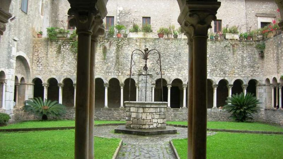 Chiostro in pietra con porticato ad archi e colonne su tre lati, affacciato su un prato centrale attraversato da un sentiero in ciottoli. Al centro si vede un pozzo in muratura con struttura in ferro, con muri in pietra e vasi di piante sul livello superiore.