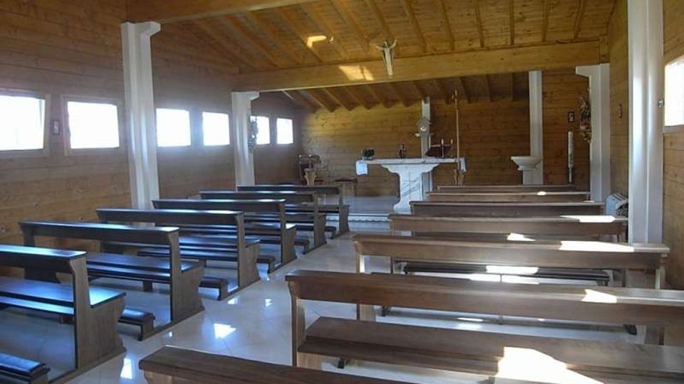 Interno di una piccola cappella in legno con soffitto a travi, file di panche scure e finestre rettangolari laterali che lasciano entrare luce naturale. In fondo si vede l’altare bianco con crocifisso, sostenuto da colonne bianche lungo la navata.