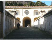 Scalinata in pietra tra due alti muri che conduce a un edificio chiaro con portico ad archi, finestre ad arco e rosone centrale sotto un tetto in coppi. Sullo sfondo si vedono alberi e un pendio collinare.