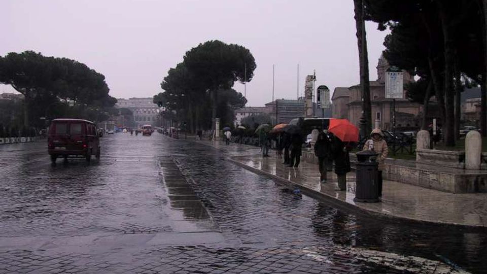 Ampio viale urbano in una giornata piovosa, con strada in pavé bagnata e riflessi sull’asfalto. Ai lati si vedono marciapiedi con alberi ad alto fusto, lampioni e alcuni edifici sullo sfondo.