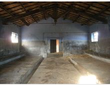Interno di una stalla o fienile in muratura con tetto a capriate in legno e pavimento in cemento, con due lunghe pedane laterali e canalette centrali. Pareti intonacate e consumate, due piccole finestre ai lati e una porta aperta sul fondo che lascia entrare luce naturale.